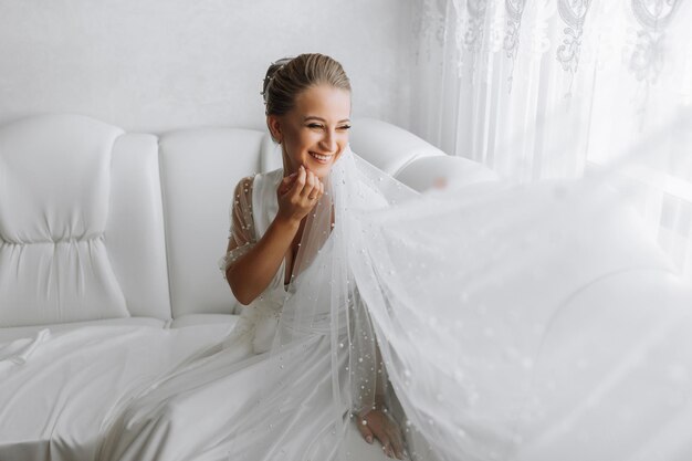 Uma noiva linda com um penteado clássico vestindo um roupão longo e um véu longo está sentada e posando em um sofá branco em seu quarto A noiva em um fundo branco
