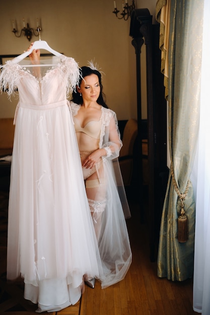 Uma noiva vestida com um vestido transparente e calcinha boudoir segura o vestido de noiva nas mãos, no interior da casa.