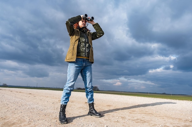 Uma ornitóloga observa pássaros com binóculos em uma estrada de cascalho contra o fundo de um céu tempestuoso