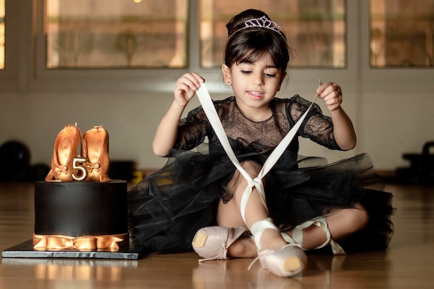 Uma pequena bailarina em um vestido de balé preto, amarrando os sapatos, ao lado de seu bolo de aniversário de cinco anos