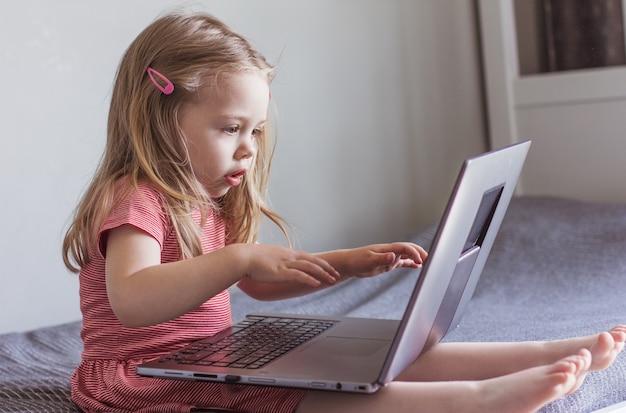 Uma pequena menina loira com um vestido vermelho se senta na cama e olha para a tela do laptop. Comunicação online, comunicação por vídeo, educação