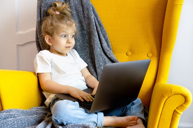 Uma pequena menina loira parece muito surpresa e olha para seu laptop enquanto está sentada em uma cadeira em casa