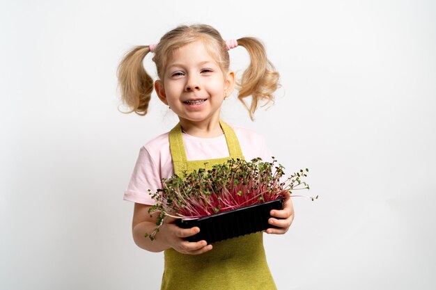 Uma pequena menina loira sorri e segura uma muda de micro-verdes nas mãos. conceito de jardinagem e plantio.
