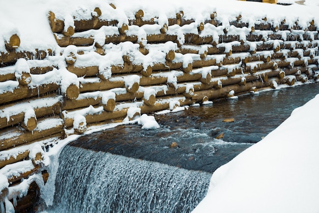 Uma pequena parede de pequenos troncos envolve um pequeno riacho na montanha com cachoeiras em uma floresta de inverno perto de árvores nuas nas montanhas dos Cárpatos