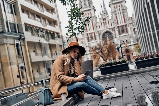 Uma pequena pausa para o café. Mulher jovem e atraente com chapéu e casaco usando seu telefone inteligente enquanto está sentado ao ar livre com a igreja ao fundo