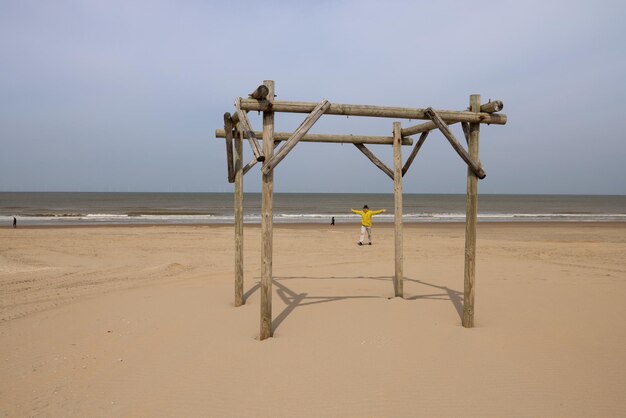 Uma pérgola de madeira solitária em uma praia de areia um homem de casaco amarelo passa pela praia