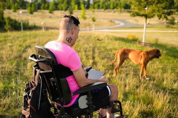 Uma pessoa com deficiência com tatuagem LGBT em cadeira de rodas passeia com seu cachorro no parque