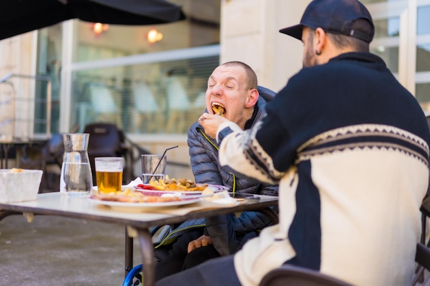 Uma pessoa com deficiência comendo no terraço de um restaurante e um amigo ajudando-o a comer