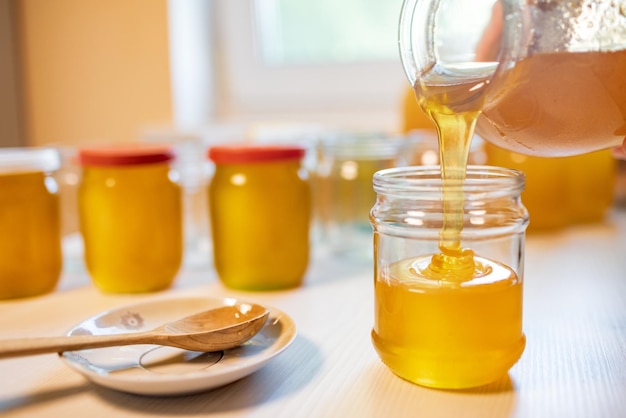 Uma pessoa desconhecida derrama mel dourado doce em potes transparentes e vazios sobre uma grande mesa de madeira branca em um dia ensolarado