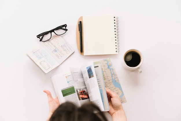 Foto uma pessoa virando as páginas do guia turístico com passaporte; óculos; bloco de notas em espiral e xícara de café em pano de fundo branco