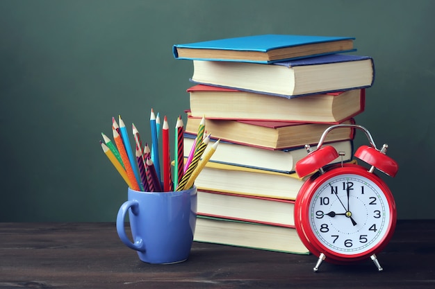 Uma pilha de livros, pastéis e despertador vermelho em cima da mesa.