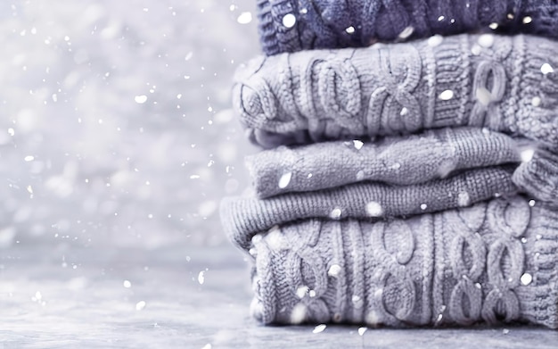 uma pilha de suéteres de inverno de malha em vários tons de cinza em fundo cinza caindo neve