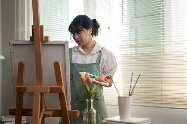 Uma pintora asiática pensativa e criativa concentra-se na pintura em uma tela em seu estúdio de arte