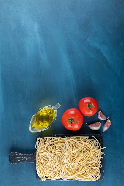 Uma placa de madeira preta de macarrão cru com dois tomates vermelhos frescos e óleo sobre uma superfície azul.