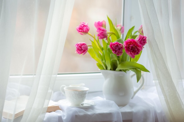 Uma primavera ainda vida um buquê de tulipas em um vaso uma xícara de chá e um livro velho estão deitados na janela