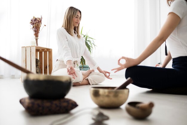 Foto uma professora de ioga e seu aluno meditam sentados um em frente ao outro há vários objetos como tigelas tibetanas uma figura de buda e flores secas