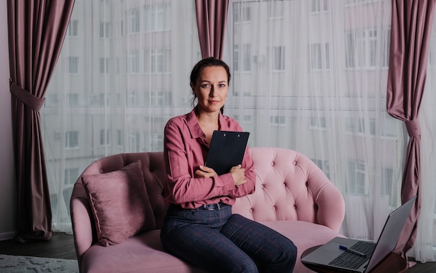 Uma psicóloga de camisa rosa está sentada no sofá com um laptop