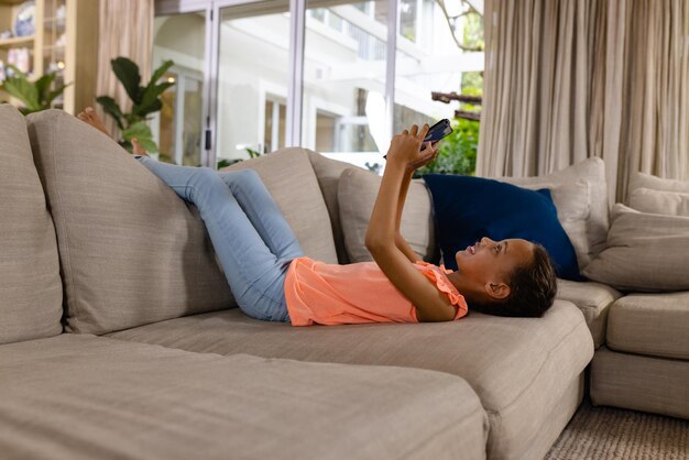 Foto uma rapariga bi-racial feliz deitada no sofá e a usar um tablet na sala de estar. infância, tempo livre, tecnologia, comunicação, entretenimento, estilo de vida e vida doméstica inalterados.