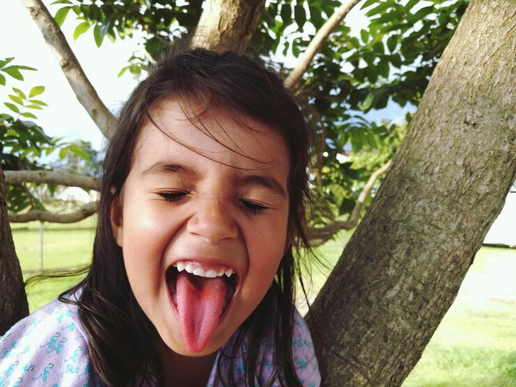 Foto uma rapariga com os olhos fechados a esticar a língua contra uma árvore no quintal.