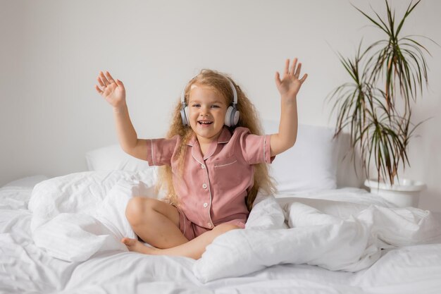 Foto uma rapariga de pijama rosa e fones de ouvido brancos diverte-se sentada na cama.