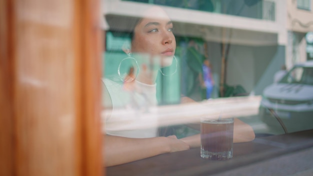 Uma rapariga sonhadora a relaxar num café com vista pela janela. Uma mulher asiática a beber.