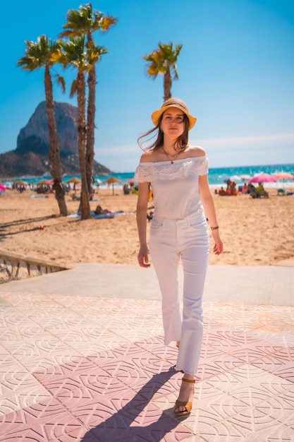 Uma ruiva caucasiana vestida de branco e com um chapéu de palha caminhando pela praia