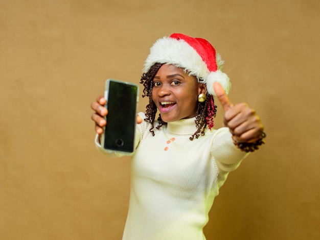 Foto uma senhora africana apontando um smartphone para a câmera, fazendo um gesto de polegar para cima e também com um boné de natal na cabeça