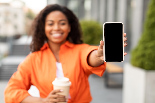 Uma senhora afro-americana milenar sorridente em smartphone casual mostrando com tela em branco desfruta de uma xícara de café