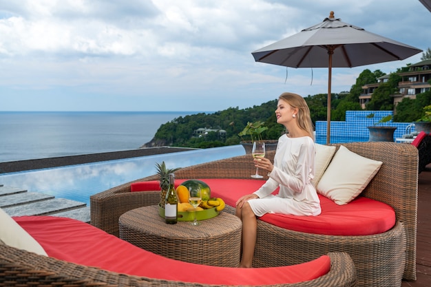 Uma senhora deslumbrante em vestido branco relaxante perto da piscina. menina sentada no sofá do rattan e bebendo vinho branco. enjoyng e relaxamento