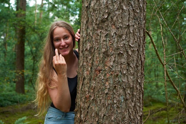 Uma senhora loira caucasiana com cabelo comprido olhando por trás de um tronco de árvore mostrando um dedo médio