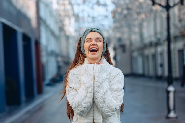 Uma sessão de fotos da cidade. Retrato de uma jovem feliz em uma roupa de malha branca com as mãos pressionadas para ela. Demonstração de prazer com os olhos fechados, boca aberta e um grande sorriso. Fundo desfocado da cidade.