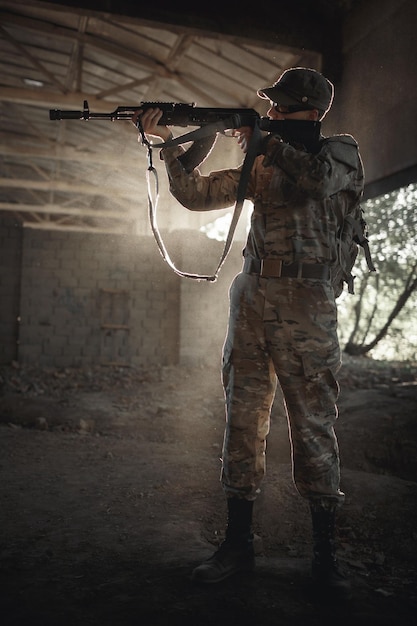 Uma silhueta de um soldado em um uniforme de proteção de camuflagem em óculos pretos táticos e um boné com uma metralhadora nas mãos fica dentro de uma grande casa permitida