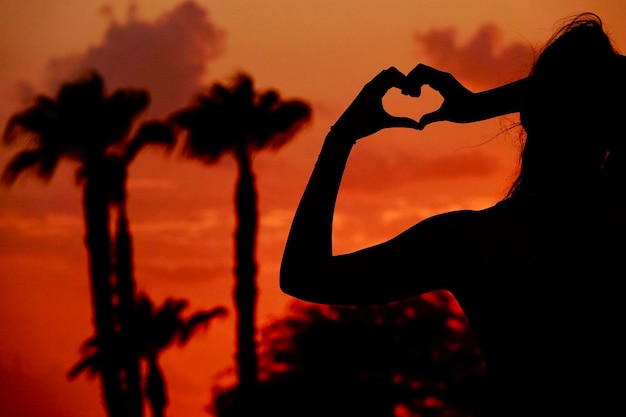 Uma silhueta de uma menina fazendo uma forma de coração com as mãos durante um pôr-do-sol de cor laranja