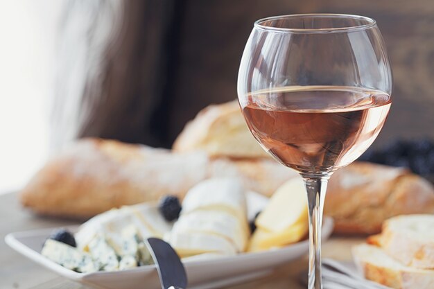 Uma taça de vinho rosé servido com prato de queijo, amoras e baguetes. Variedade de queijo com frutas em fundo de madeira.