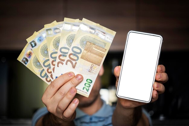 Uma tela branca vazia em um smartphone para anunciar o aplicativo, o homem usa roupas casuais, camisa azul na mão, leque de dinheiro em notas de euro, usa celular