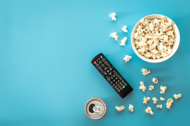 Uma tigela de pipoca um controle remoto de TV uma lata com uma bebida em um fundo azul