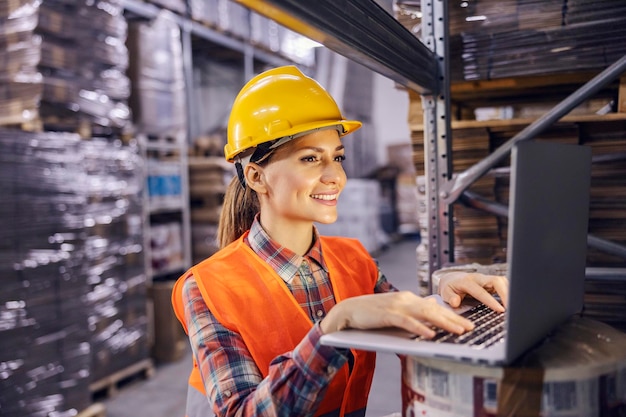 Uma trabalhadora de depósito com capacete digitando e inserindo dados em seu laptop enquanto sorri para ele