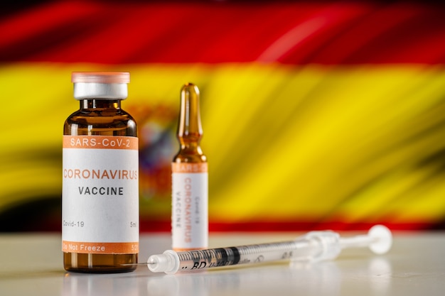 Uma vacina contra o coronavírus COVID-19 e uma seringa médica no fundo da bandeira espanhola. O conceito de tratamento, imunização e vacinação em caso de pandemia na Espanha.