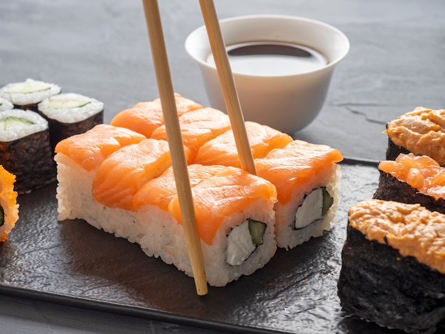 Uma variedade de rolos japoneses e sushi em uma placa preta texturizada. vista lateral. pauzinhos de bambu segurando um único rolo