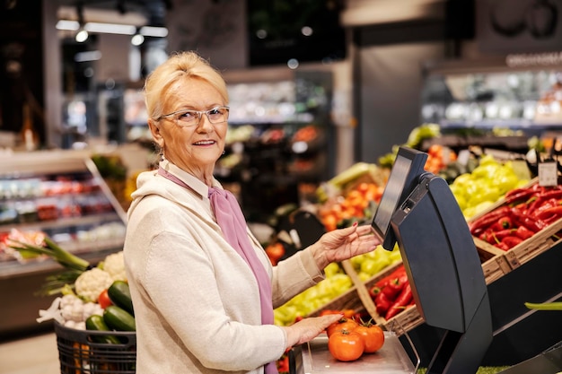 Uma velha alegre está medindo tomates em escalas no supermercado e sorrindo para a câmera