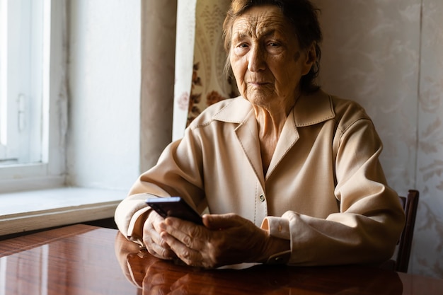 Uma velha avó caucasiana sênior com rugas profundas está sentada em casa, usa seu telefone inteligente na câmera frontal do braço estendido para uma videochamada.