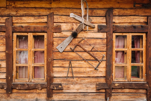 Uma velha parede de madeira de uma casa com janelas na aldeia e ferramentas penduradas