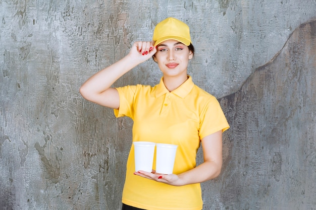 Uma vendedora de uniforme amarelo segurando dois copos plásticos com bebida.