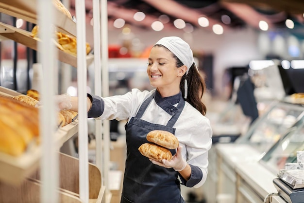 Uma vendedora feliz do departamento de padaria organiza massa fresca nas prateleiras do supermercado