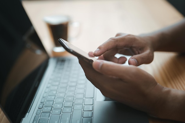 Uma visão aproximada das mãos masculinas tocando um homem de telefone celular usando seu smartphone enquanto está sentado na mesa de madeira