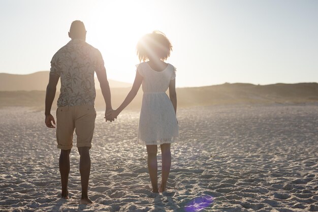 Uma visão traseira de um casal de raça mista aproveitando o tempo livre na praia em um dia ensolarado juntos, de mãos dadas com o sol brilhando em seus rostos.