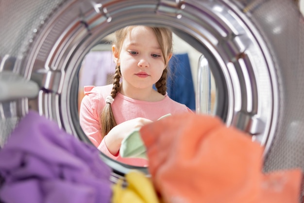 Uma vista de dentro do tambor para a lavanderia, uma garotinha bonitinha está colocando roupas coloridas