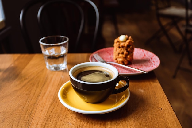 Uma xícara cerâmica de café preto uma sobremesa servida em um prato e um copo de água na mesa de madeira em um café