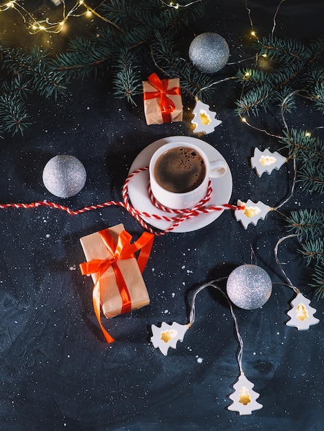 Uma xícara de café branca, presentes de Natal e uma guirlanda em forma de árvore de Natal em uma mesa escura à noite. Atmosfera caseira de férias