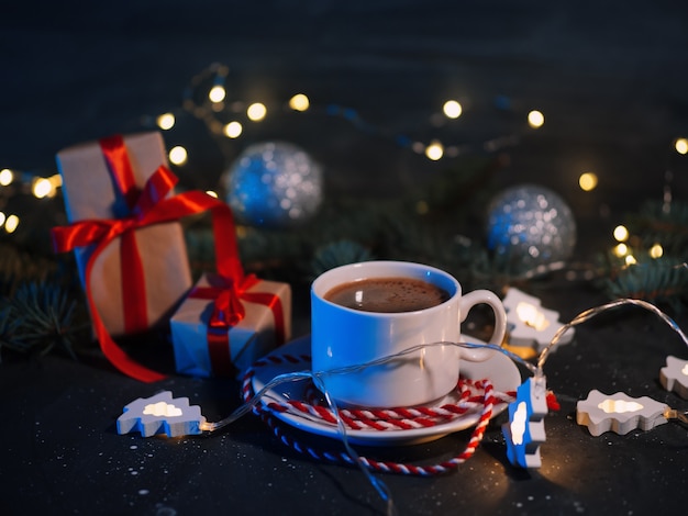 Uma xícara de café branca, presentes de natal e uma guirlanda em forma de árvore de natal em uma mesa escura à noite. atmosfera caseira de férias